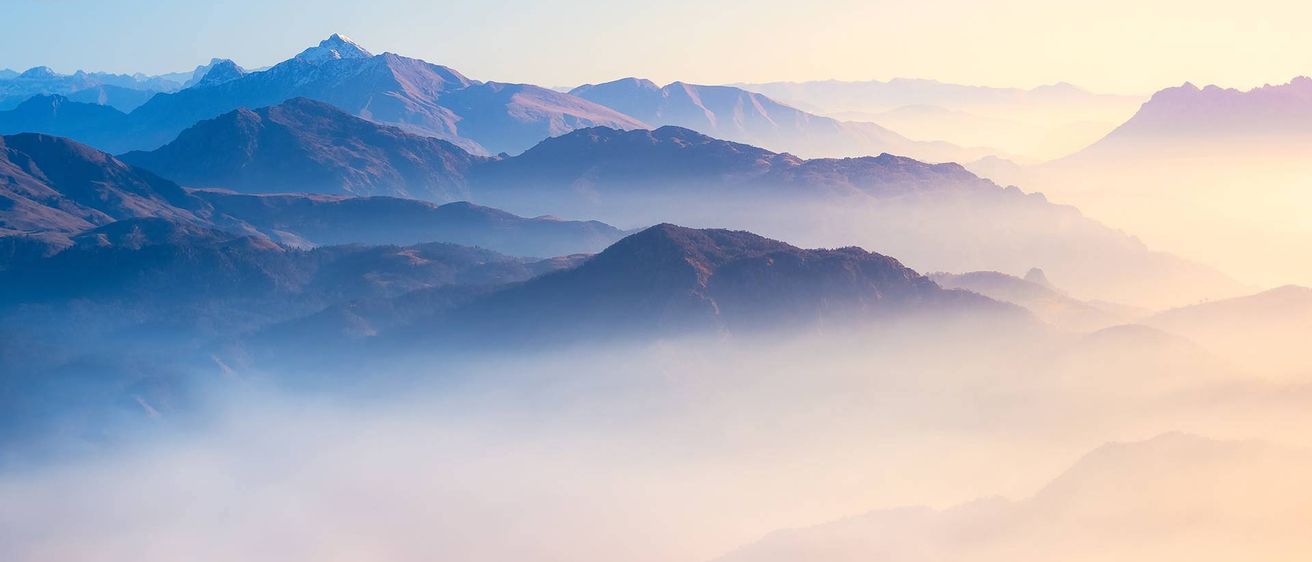 Eine neblige Morgenszene mit einer Bergkette in der Ferne. Die Gipfel sind in Nebel gehüllt, und der Himmel ist ein sanfter Farbverlauf von Rosa und Blau.