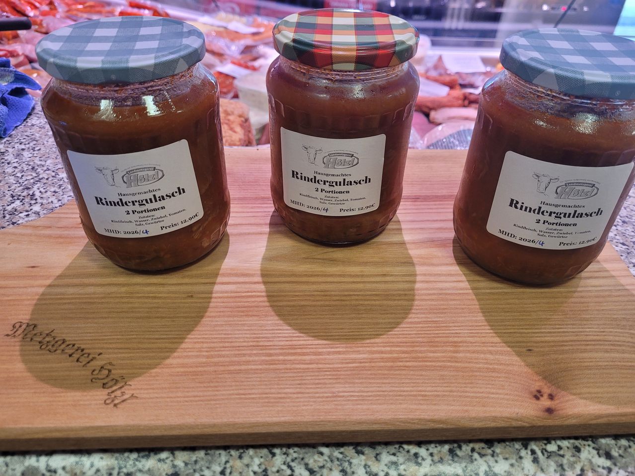 Three jars of home-made beef stew sit on a wooden board. Each jar has a label with the brand name and date of production. The jars have checkered lids.