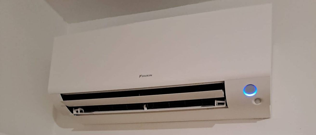 Eine weiße Klimaanlage, die an der Decke über einer Tür montiert ist, die zu einem Raum mit Holzboden und einem Schreibtisch führt.