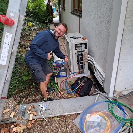 Ein Mann in einer blauen Jacke arbeitet an einer Klimaanlage, lächelt und gibt einen Daumen hoch.