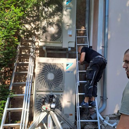 Ein Mann klettert eine Leiter und repariert eine Klimaanlage. Er trägt ein schwarzes Shirt und Hose. Ein Mann in einem grünen Shirt steht neben ihm.