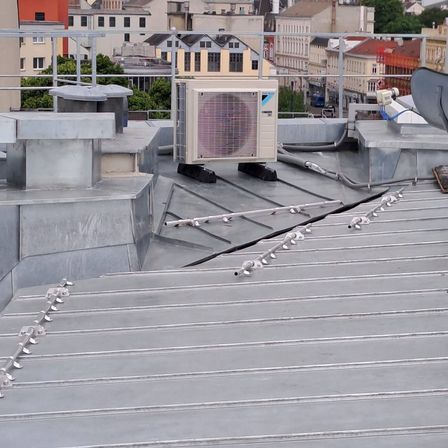 Ein Blick auf ein städtisches Dach zeigt Metallstrukturen, eine Daikin-Klimaanlage und verschiedene Rohre und Kabel. Das Dach ist grau, und im Hintergrund befinden sich Gebäude.