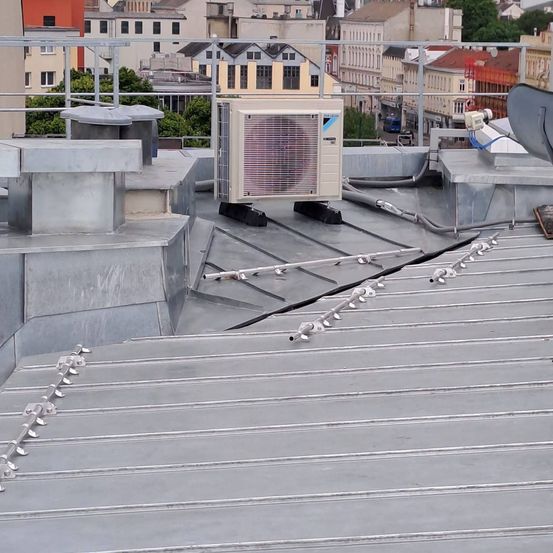 Ein Blick auf ein städtisches Dach zeigt Metallstrukturen, eine Daikin-Klimaanlage und verschiedene Rohre und Kabel. Das Dach ist grau, und im Hintergrund befinden sich Gebäude.
