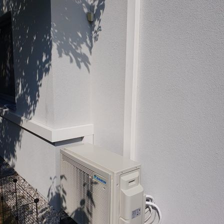 Ein weißes Daikin-Klimagerät ist an der Wand eines weißen Hauses montiert. Ein Schatten eines Baumes fällt auf die Wand.