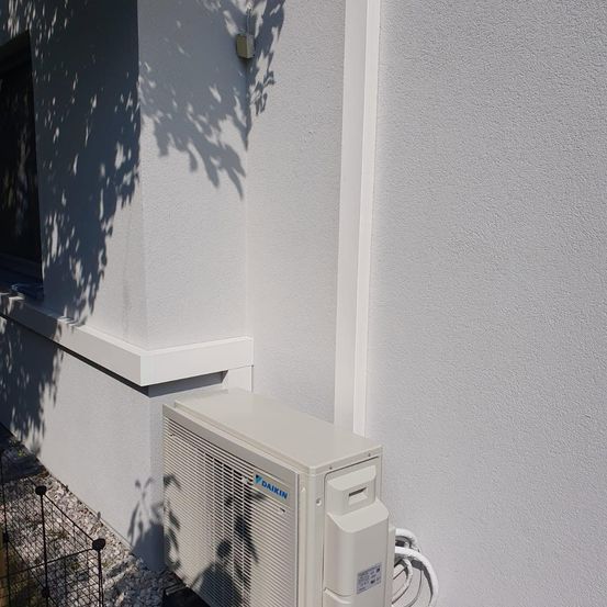 Ein weißes Daikin-Klimagerät ist an der Wand eines weißen Hauses montiert. Ein Schatten eines Baumes fällt auf die Wand.
