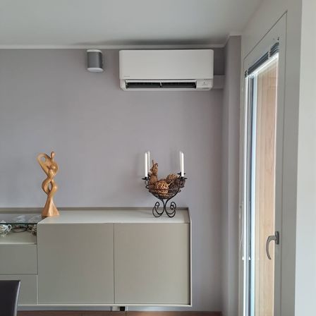 Ein Raum mit einem weißen Schrank, grauen Wänden und einer weißen Klimaanlage. Eine Klimaanlage ist an der Wand montiert. Ein Glastisch hat eine Figurine und einen Kerzenhalter. Eine Glastür führt nach draußen.