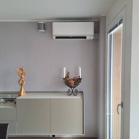 Ein Raum mit einem weißen Schrank, grauen Wänden und einer weißen Klimaanlage. Eine Klimaanlage ist an der Wand montiert. Ein Glastisch hat eine Figurine und einen Kerzenhalter. Eine Glastür führt nach draußen.