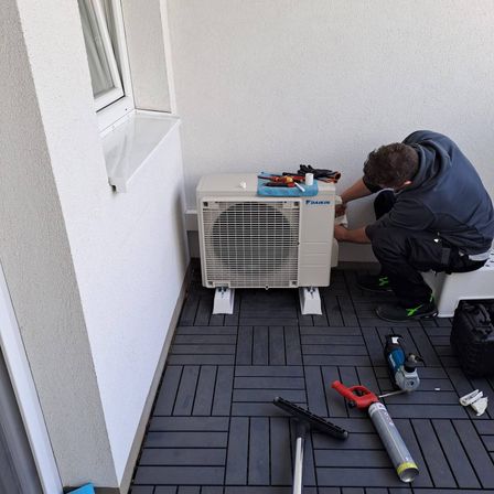 Ein Mann in einer blauen Jacke arbeitet auf einem Balkon an einer Klimaanlage. Verschiedene Werkzeuge sind um ihn herum verstreut.