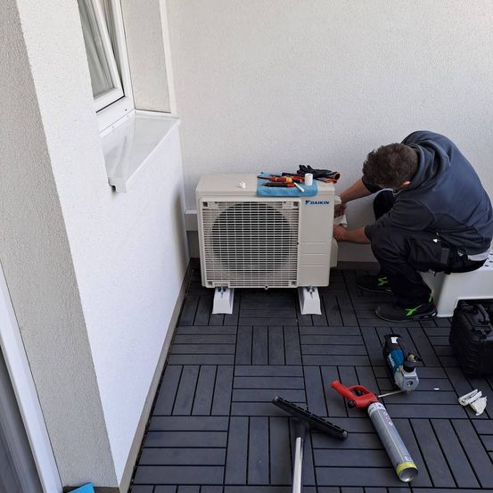 Ein Mann in einer blauen Jacke arbeitet auf einem Balkon an einer Klimaanlage. Verschiedene Werkzeuge sind um ihn herum verstreut.