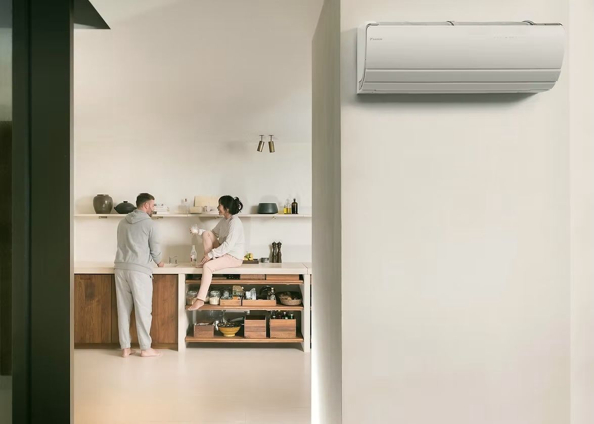 Ein Mann und eine Frau befinden sich in einer modernen Küche, die Frau sitzt auf einem Tresen, während der Mann in der Nähe steht. Sie sind beide barfuß. Die Küche hat weiße Wände, Holzschränke und einen weißen Fliesenboden. An der Wand befindet sich eine Klimaanlage.