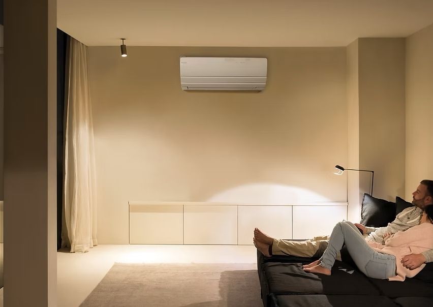 Ein Wohnzimmer mit einem Sofa und zwei darauf liegenden Personen. Eine Deckenlampe und eine Klimaanlage sind an der Wand montiert.