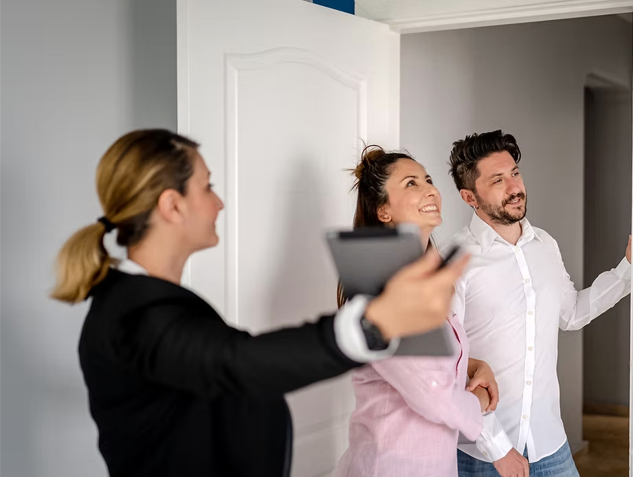 A woman in a black suit points to a tablet in her hand while a smiling woman and man stand by the open door.