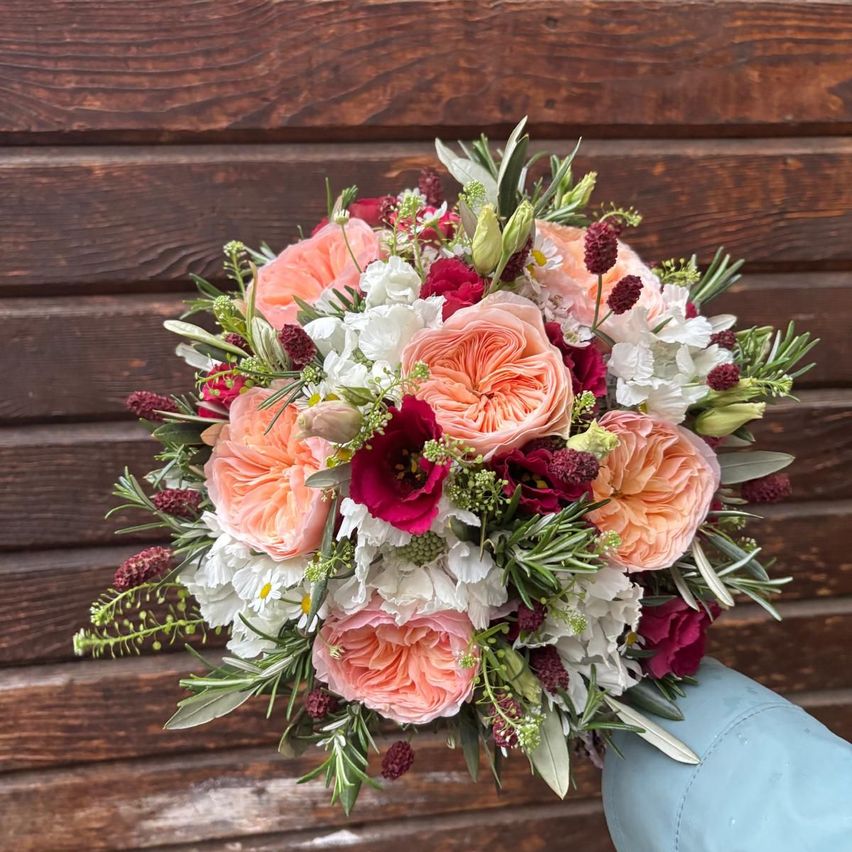 Ein Blumenstrauß mit rosa, weißen und roten Rosen sowie Grünpflanzen, gehalten in einer blauen Hand vor einem hölzernen Hintergrund.