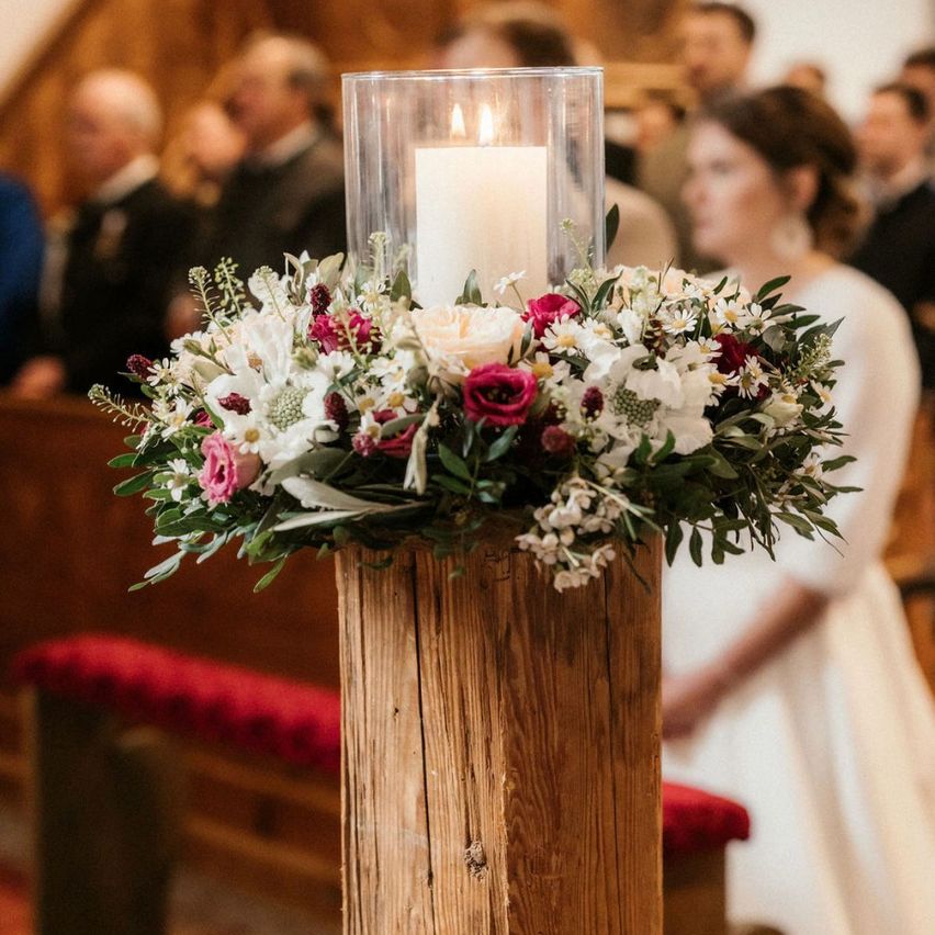 Eine Hochzeitszeremonie mit einem hölzernen Pfeiler, der eine hohe Kerze umgibt, umgeben von einer Blumenarrangement, eine Braut steht im Hintergrund.