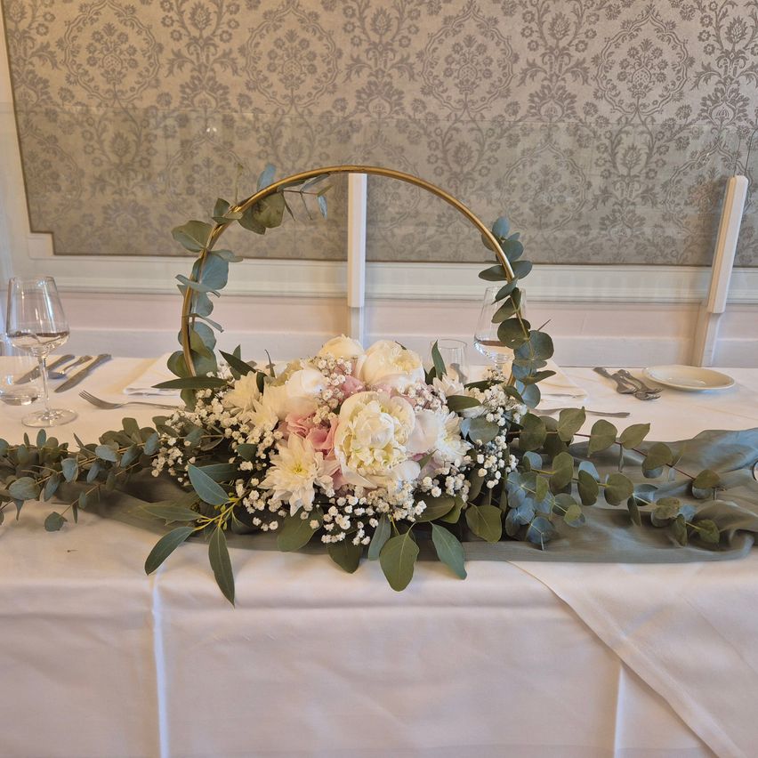 Eine weiße Tischdecke zeigt einen Blumenstrauss mit rosa und weißen Blumen in einem goldenen Korb, umgeben von grünen Blättern, mit Weingläsern und Besteck auf dem Tisch.