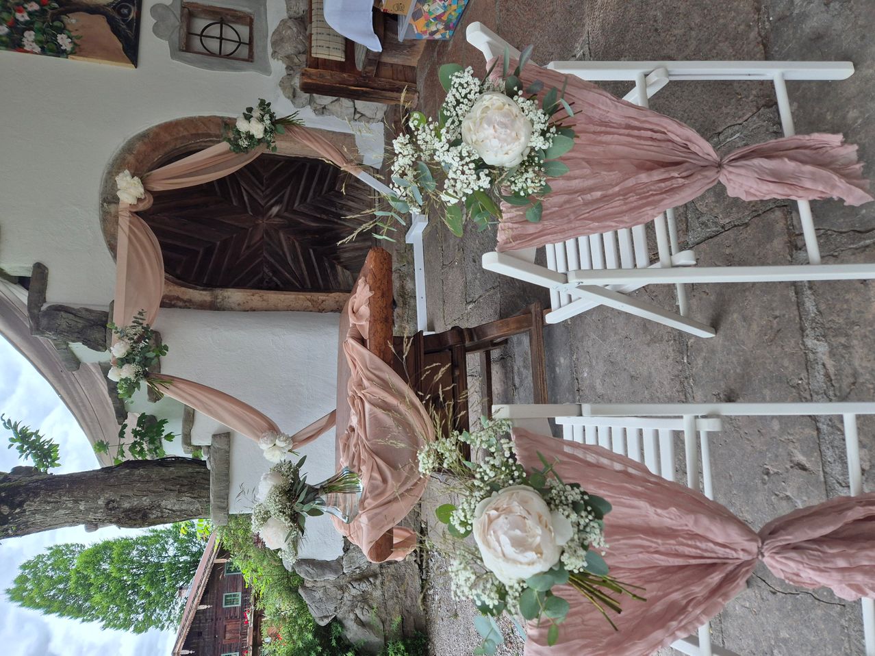 Eine rustikale Zeremonie-Aufstellung mit Holzstühlen, die mit rosa Stoff und Blumenarrangements geschmückt sind, vor einem Hintergrund aus einer Steinmauer und einem gewölbten Türbogen.