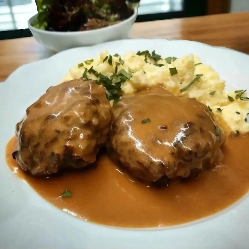 A white plate holds two meatballs in gravy, accompanied by mashed potatoes and a salad in a small bowl on a wooden table.