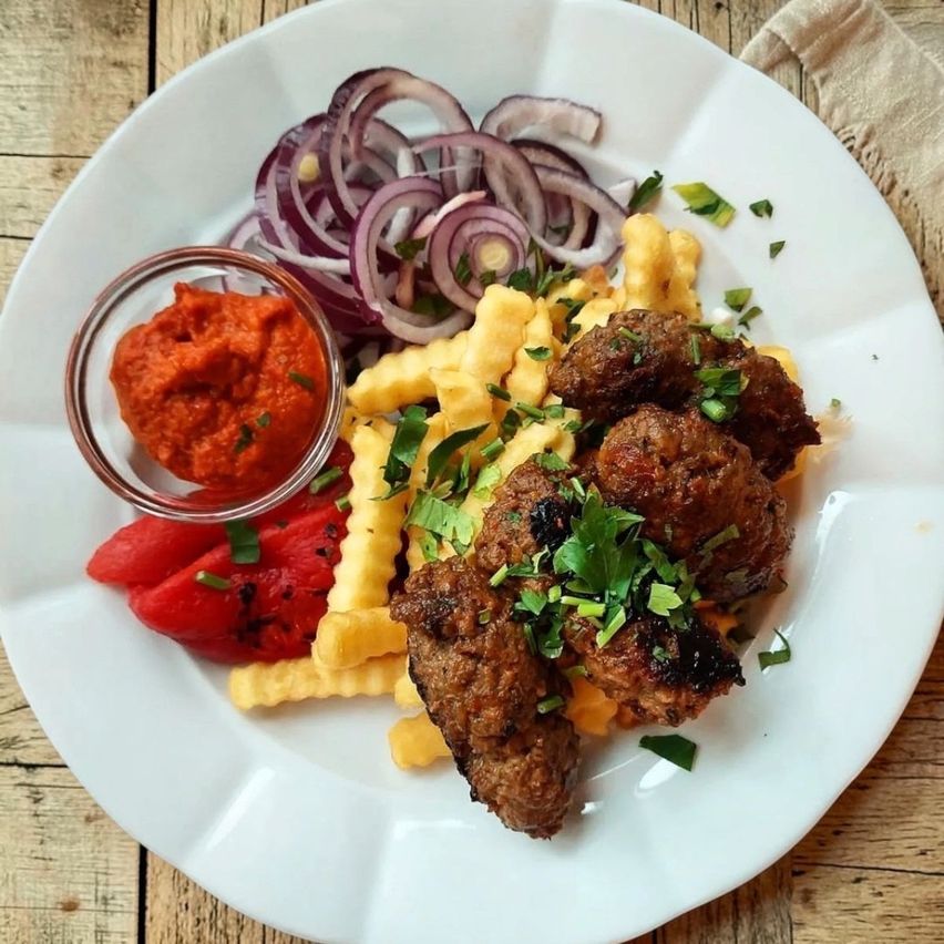 A white plate with grilled meatballs, French fries, red onions, and a small bowl of red sauce.