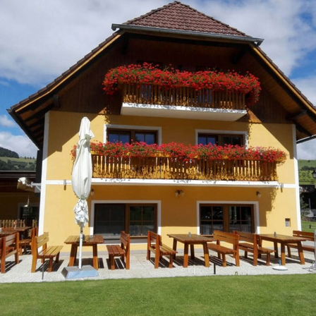 Ein gelbes Haus mit roten Blumen auf den Balkonen, Holzbänken und Tischen draußen. Das Haus hat ein braunes Dach und ist von einer Rasenfläche umgeben.