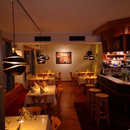 Ein Innenrestaurant mit mehreren für das Essen gedeckten Tischen. Holzstühle umgeben die Tische, und dekorative Lampen hängen von der Decke. Der Raum hat Holzböden und weiße Wände, mit einer Bar an der rechten Seite.
