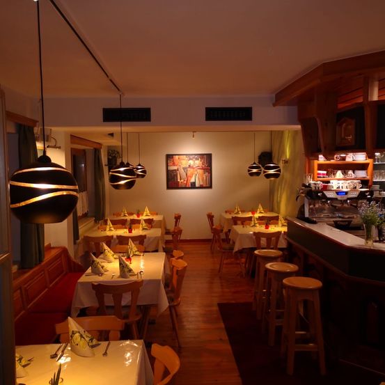 Ein Innenrestaurant mit mehreren für das Essen gedeckten Tischen. Holzstühle umgeben die Tische, und dekorative Lampen hängen von der Decke. Der Raum hat Holzböden und weiße Wände, mit einer Bar an der rechten Seite.