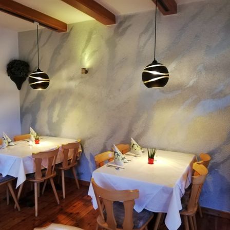 Ein Restaurantinnenraum mit zwei in weiße Tücher gehüllten Tischen, die jeweils mit einer Pflanze und einer Serviette geschmückt sind. Holzstühle umgeben die Tische, und zwei Pendelleuchten hängen von der Decke. Die Wände haben eine strukturierte Oberfläche.