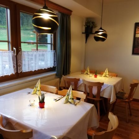 Ein Essbereich mit zwei Tischen, jeder mit einer Pflanze, Kerzen und gefalteten Servietten bestückt. Der Raum hat ein Fenster mit Vorhängen, einem Heizkörper und zwei Lampen, die von der Decke hängen.