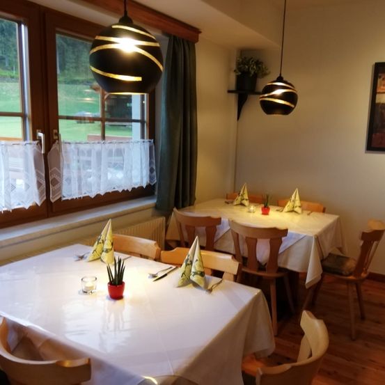 Ein Essbereich mit zwei Tischen, jeder mit einer Pflanze, Kerzen und gefalteten Servietten bestückt. Der Raum hat ein Fenster mit Vorhängen, einem Heizkörper und zwei Lampen, die von der Decke hängen.