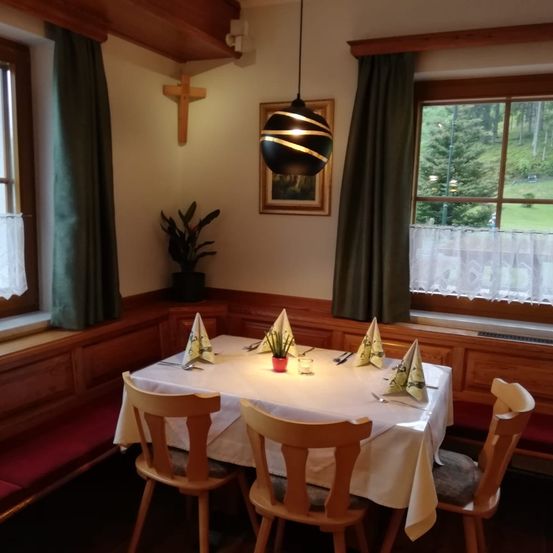 Ein Essbereich mit zwei Stühlen für eine Mahlzeit. Ein Tisch hält eine Topfpflanze, Besteck und Servietten. Ein Kreuz ist an der Wand über einem gerahmten Bild, und eine Lampe hängt über dem Tisch. Draußen gibt es eine Aussicht auf Bäume.