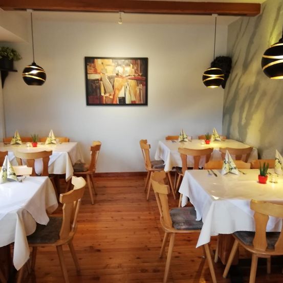 Ein leerer Essbereich mit Holzböden und weißen Tischdecken. Einige Tische und Stühle sind aufgestellt, und dekorative Pflanzen stehen auf den Tischen. Ein gerahmtes Bild ist an der Wand über den Tischen montiert. Die Wand hat ein grau-weißes Design.