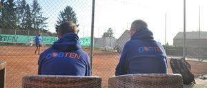 Zwei Männer in blauen Sweatshirts mit der Aufschrift 'DOBTEN' sitzen auf Korbstühlen auf einem Tennisplatz und beobachten einen Spieler auf dem Platz. Ein Zaun umgibt den Platz, und ein Tennisschläger liegt in der Nähe.