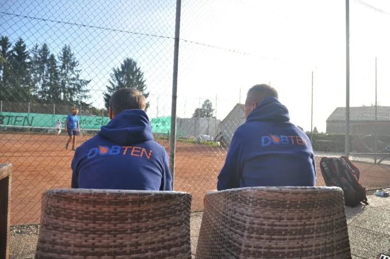 Zwei Männer in blauen Sweatshirts mit der Aufschrift 'DOBTEN' sitzen auf Korbstühlen auf einem Tennisplatz und beobachten einen Spieler auf dem Platz. Ein Zaun umgibt den Platz, und ein Tennisschläger liegt in der Nähe.