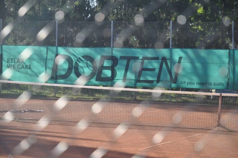 Ein Tennisplatz mit einem Banner, auf dem 'DOBTEN' und der Slogan 'Entspannen Sie sich, wir kümmern uns. Alles, was Sie brauchen, sind Bälle' steht. Der Platz ist von einem Zaun umgeben, mit Bäumen im Hintergrund.
