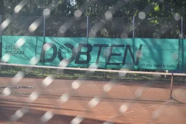 Ein Tennisplatz ist eingezäunt. Ein grünes Banner mit dem Wort 'Dobten' ist an der Umzäunung befestigt. Ein Netz ist vor dem Platz aufgestellt.