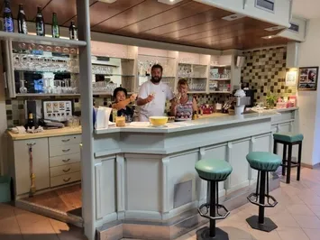 Eine Bar mit einer Theke und Hockern, an der drei Personen stehen und lächeln, mit Regalen mit Gläsern und Flaschen im Hintergrund.