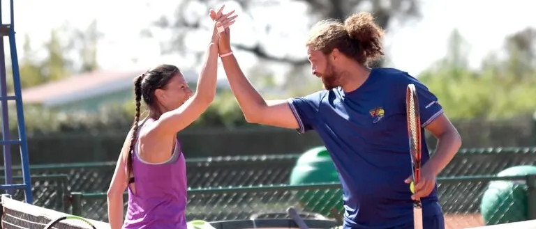 Ein Mann und eine Frau stehen auf einem Tennisplatz. Die Frau hält einen Tennisschläger und hat ihre Hände erhoben, und der Mann erhebt seine Hände. Sie lächeln beide.