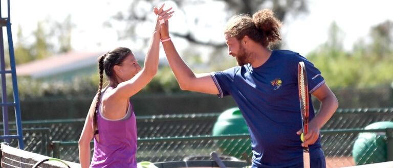 Ein Mann und eine Frau stehen auf einem Tennisplatz. Die Frau hält einen Tennisschläger und hat ihre Hände erhoben, und der Mann erhebt seine Hände. Sie lächeln beide.
