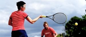 Eine Frau mit Brille schwingt einen Tennisschläger, um einen Ball zu treffen, während ein Mann vor einem Tennisballhalter auf einem Tennisplatz unter einem bewölkten Himmel steht.