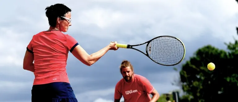Eine Frau mit Brille schwingt einen Tennisschläger, um einen Ball zu treffen, während ein Mann vor einem Tennisballhalter auf einem Tennisplatz unter einem bewölkten Himmel steht.
