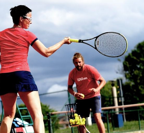Eine Frau in einem roten Shirt und schwarzen Brille schwingt einen Tennisschläger an einem sonnigen Tag, während ein Mann in einem roten Shirt mit einem Korb voller Tennisbälle hinter ihr steht.