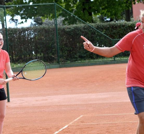 Eine Frau und ein Mann sind auf einem Tennisplatz. Die Frau hält einen Tennisschläger, während der Mann mit dem Finger auf sie zeigt.
