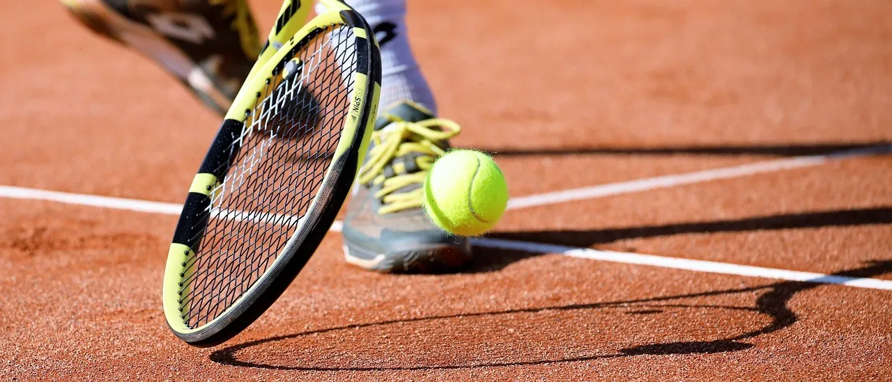 Der Fuß und Schläger eines Tennisspielers sind auf einem Sandplatz zu sehen, mit einem Tennisball in der Nähe des Fußes des Spielers.