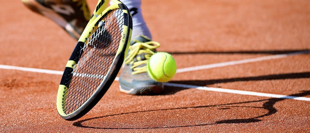 Der Fuß und Schläger eines Tennisspielers sind auf einem Sandplatz zu sehen, mit einem Tennisball in der Nähe des Fußes des Spielers.