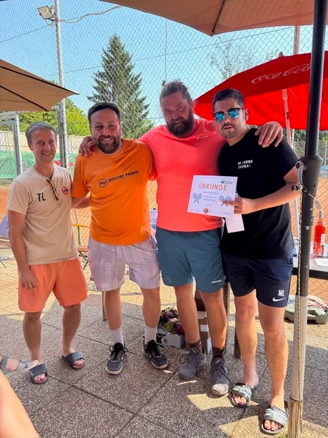 Vier Männer stehen bei einer Outdoor-Veranstaltung zusammen. Einer hält eine Urkunde, und sie alle lächeln, was auf eine Feier hindeutet. Sie befinden sich in der Nähe eines roten Regenschirms und einer Coca-Cola-Flasche.