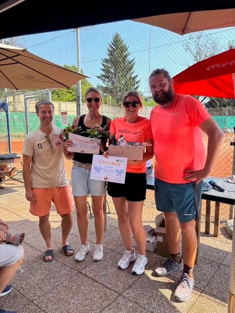Fünf Personen, zwei Frauen und drei Männer, stehen lächelnd auf einem Tennisplatz. Eine Frau hält einen Blumenstrauß und ein Zertifikat, und eine andere hält eine Schachtel. Sie stehen unter Sonnenschirmen.