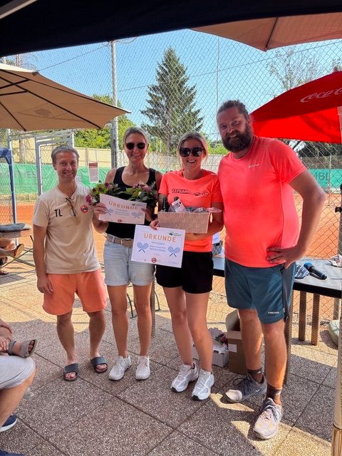 Fünf Personen, zwei Frauen und drei Männer, stehen lächelnd auf einem Tennisplatz. Eine Frau hält einen Blumenstrauß und ein Zertifikat, und eine andere hält eine Schachtel. Sie stehen unter Sonnenschirmen.