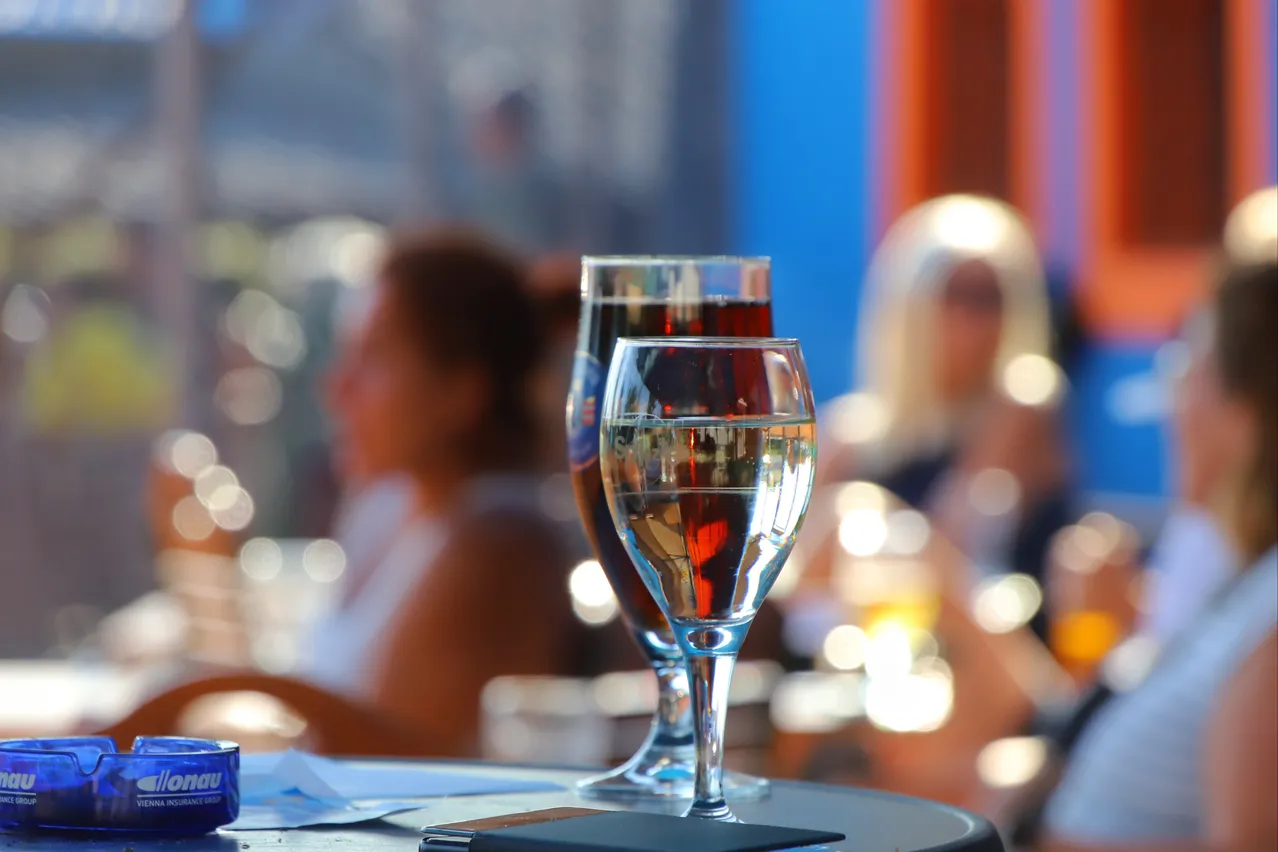 Zwei mit Flüssigkeit gefüllte Weingläser stehen auf einem Tisch, mit verschwommenen Personen im Hintergrund. Das Glas auf der linken Seite ist dunkler, möglicherweise enthält es Rotwein. Das Glas auf der rechten Seite ist klarer, wahrscheinlich enthält es Weißwein.