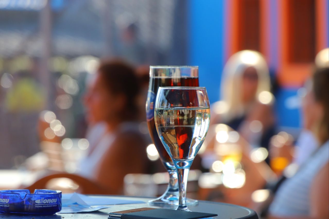 Zwei mit Flüssigkeit gefüllte Weingläser stehen auf einem Tisch, mit verschwommenen Personen im Hintergrund. Das Glas auf der linken Seite ist dunkler, möglicherweise enthält es Rotwein. Das Glas auf der rechten Seite ist klarer, wahrscheinlich enthält es Weißwein.
