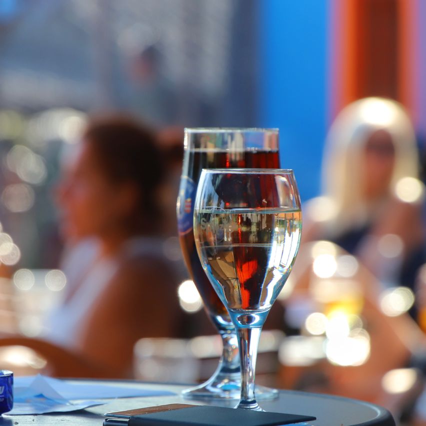 Zwei mit Flüssigkeit gefüllte Weingläser stehen auf einem Tisch, mit verschwommenen Personen im Hintergrund. Das Glas auf der linken Seite ist dunkler, möglicherweise enthält es Rotwein. Das Glas auf der rechten Seite ist klarer, wahrscheinlich enthält es Weißwein.