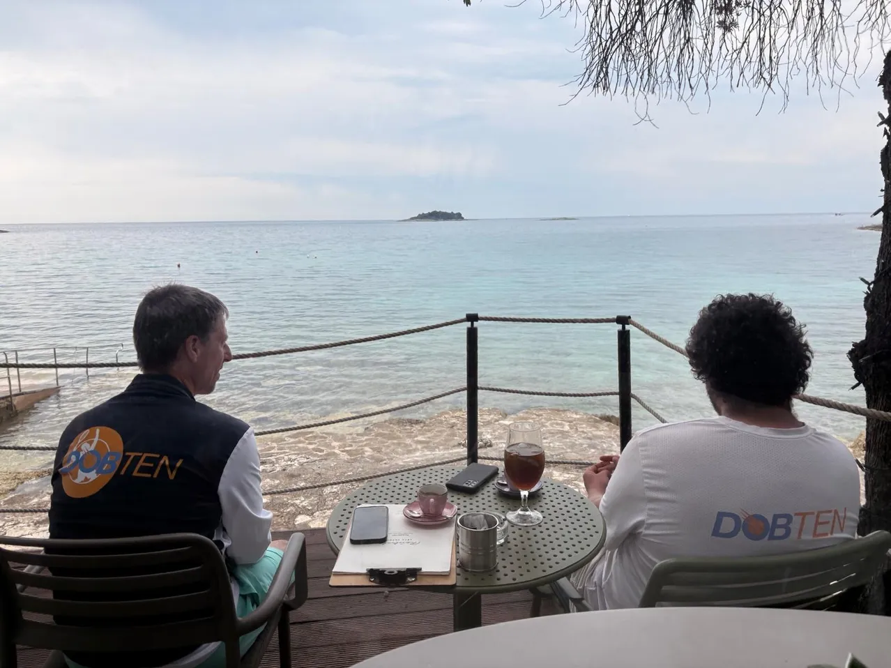 Zwei Personen sitzen auf einem Balkon mit Meerblick, eine trägt eine Weste und die andere ein weißes T-Shirt. Ein Tisch zwischen ihnen hat ein Glas, eine Tasse, ein Handy und ein Notizbuch. In der Ferne ist eine kleine Insel zu sehen.